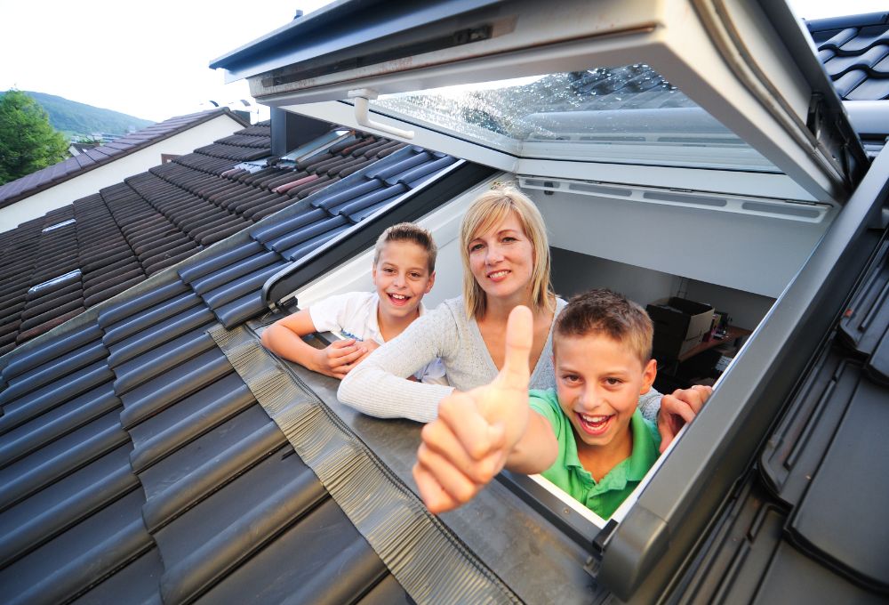 Glückliche Familie schaut aus Dachflächenfenster nach gelungener Arbeit der Dachdecker und Spengler in Leoben, Steiermark.